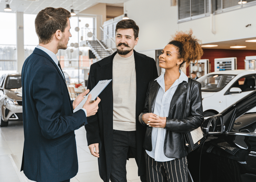 Image header for blog post A car salesman holding a clipboard talking to 2 customers about getting out of their current car loan, in a show room